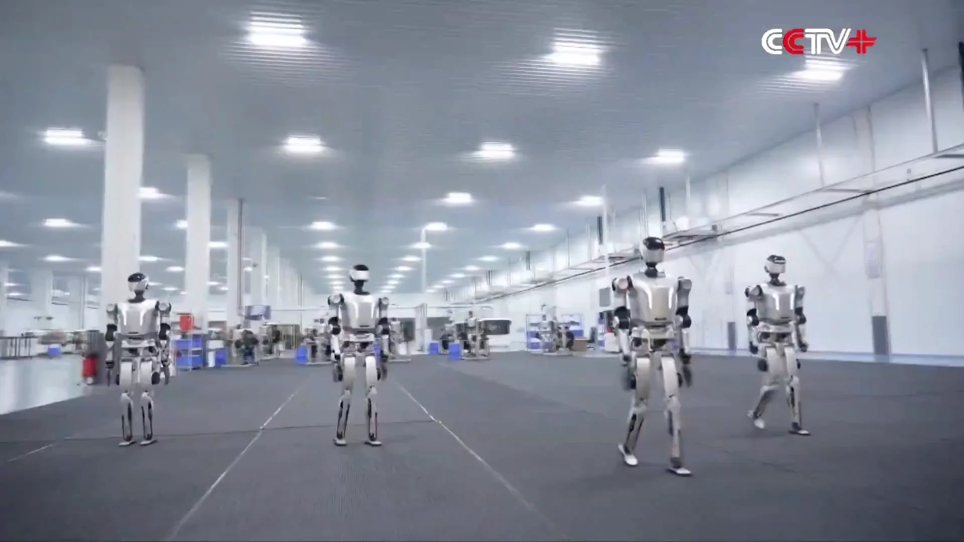 Four silver and black humanoid robots walk in formation across a gray floor inside a vast, brightly lit industrial facility with white pillars and high ceilings. A "CCTV+" logo is visible in the upper right corner.