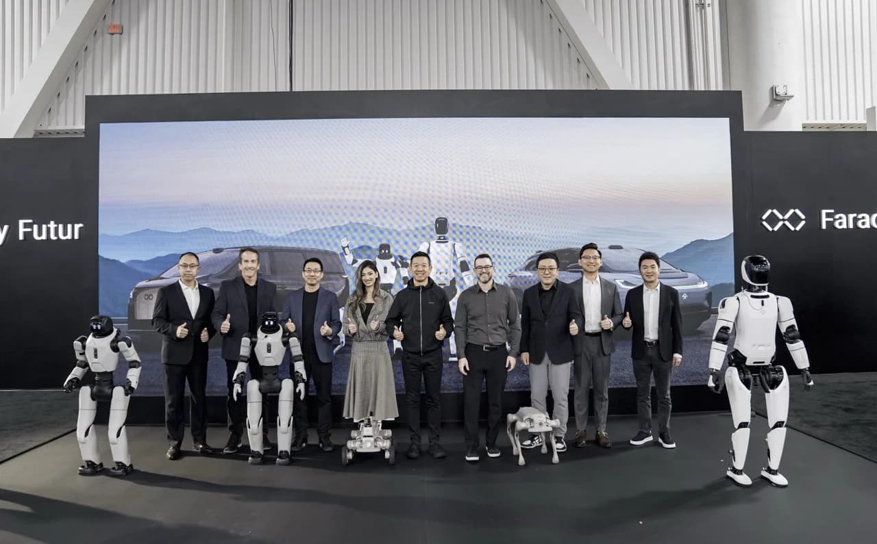 A group of nine people, including Faraday Future executives and partners, stand on a stage giving thumbs-up gestures. They are flanked by three humanoid robots and a quadruped robot, with a large digital screen behind them displaying Faraday Future branding.