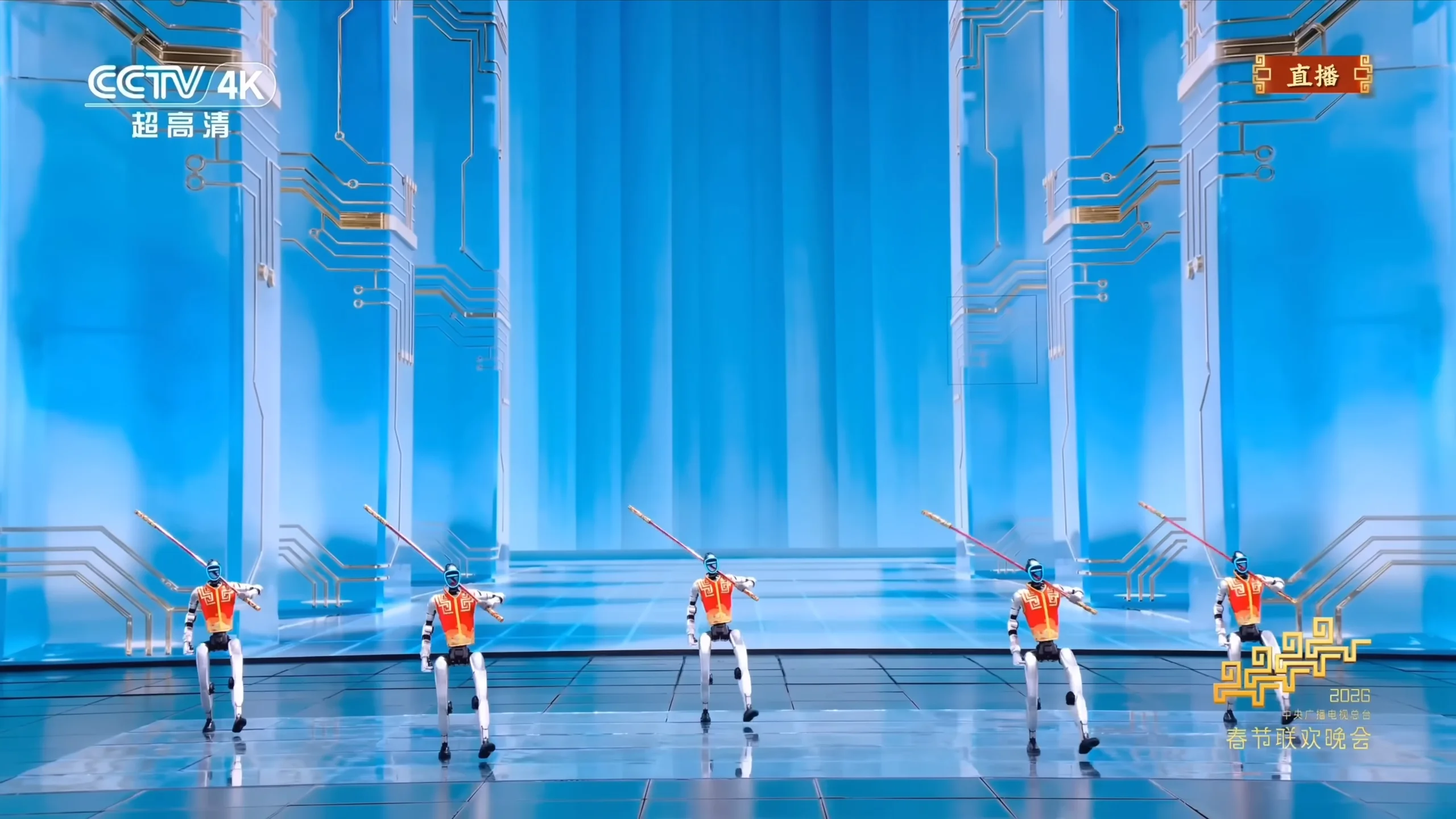 Five Unitree G1 humanoid robots stand in a row on a reflective stage during the 2026 Spring Festival Gala. Each robot wears a traditional red and orange vest and holds a long staff across its shoulders. The background features futuristic blue pillars with gold circuit-board patterns and the CCTV 4K logo.