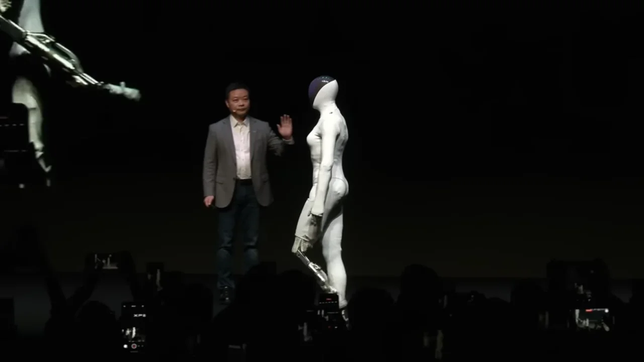 A female-styled Xpeng Iron humanoid robot walking on stage. The robot's left leg is covered in a white bodysuit, while the right leg has been stripped of its outer fabric and foam padding to reveal the metal mechanical skeleton and actuators underneath.