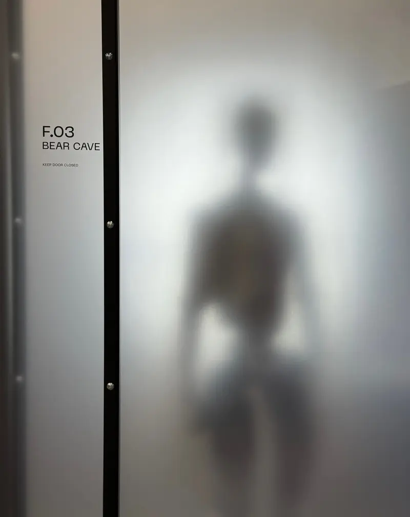 An image posted by Figure AIs CEO Brett Adcock teasing the reveal of their F.03 robot model. It stands behind a frosted glass wall so one cannot make out any details. A sign on the wall says F.03 BEAR CAVE Keep door closed.