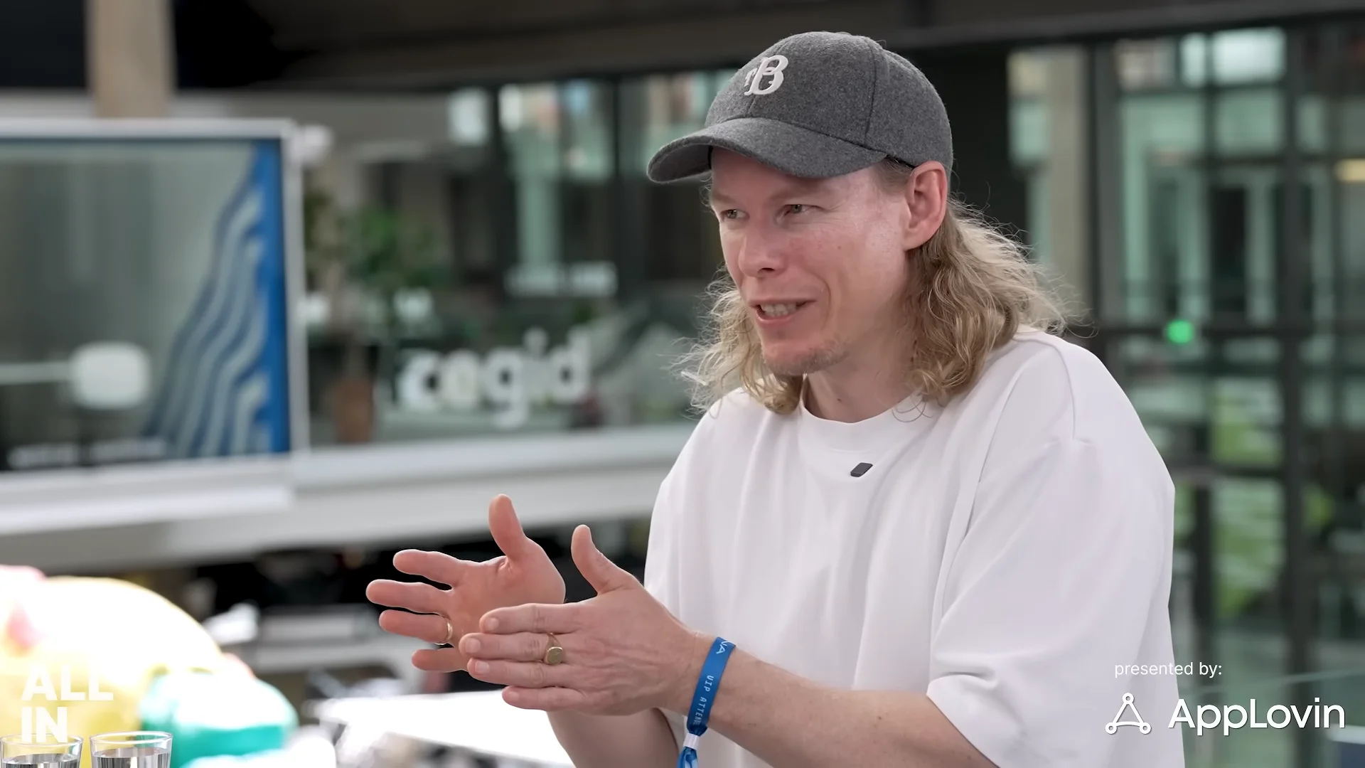 1X Founder and CEO Bernt Børnich wearing a grey baseball cap and white t-shirt, gesturing with his hands during an interview on the All-In Podcast.