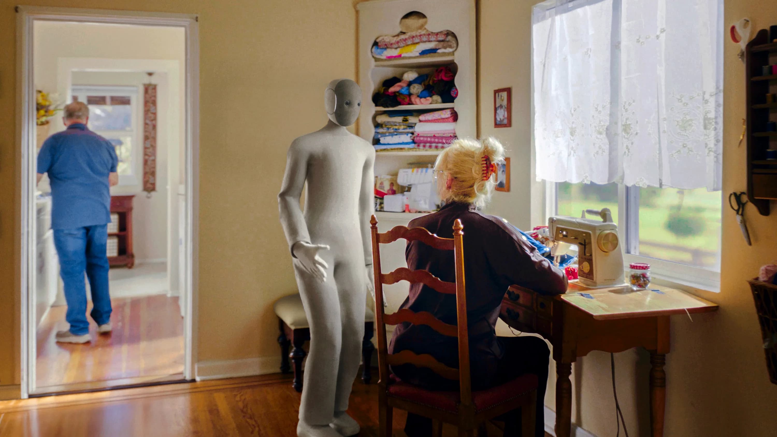 A soft-bodied, grey textile-clad 1X NEO humanoid robot standing beside an elderly woman sitting at a desk sewing machine in a sunlit living room.