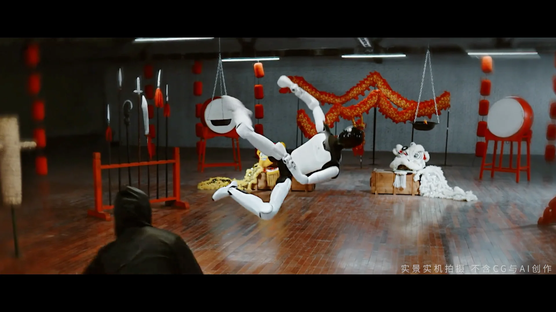 A wide-angle screenshot from a training studio showing the AgiBot Expedition A3 humanoid robot mid-air. The robot, with a white torso and black joints, is performing a horizontal flying kick. In the background, traditional Chinese martial arts equipment is visible, including red drums, weapon racks with spears, and a decorative dragon hanging from the ceiling. A person in dark clothing is seen in the lower-left foreground, watching the robot. A Chinese watermark in the bottom-right corner specifies the footage is real and CGI-free.