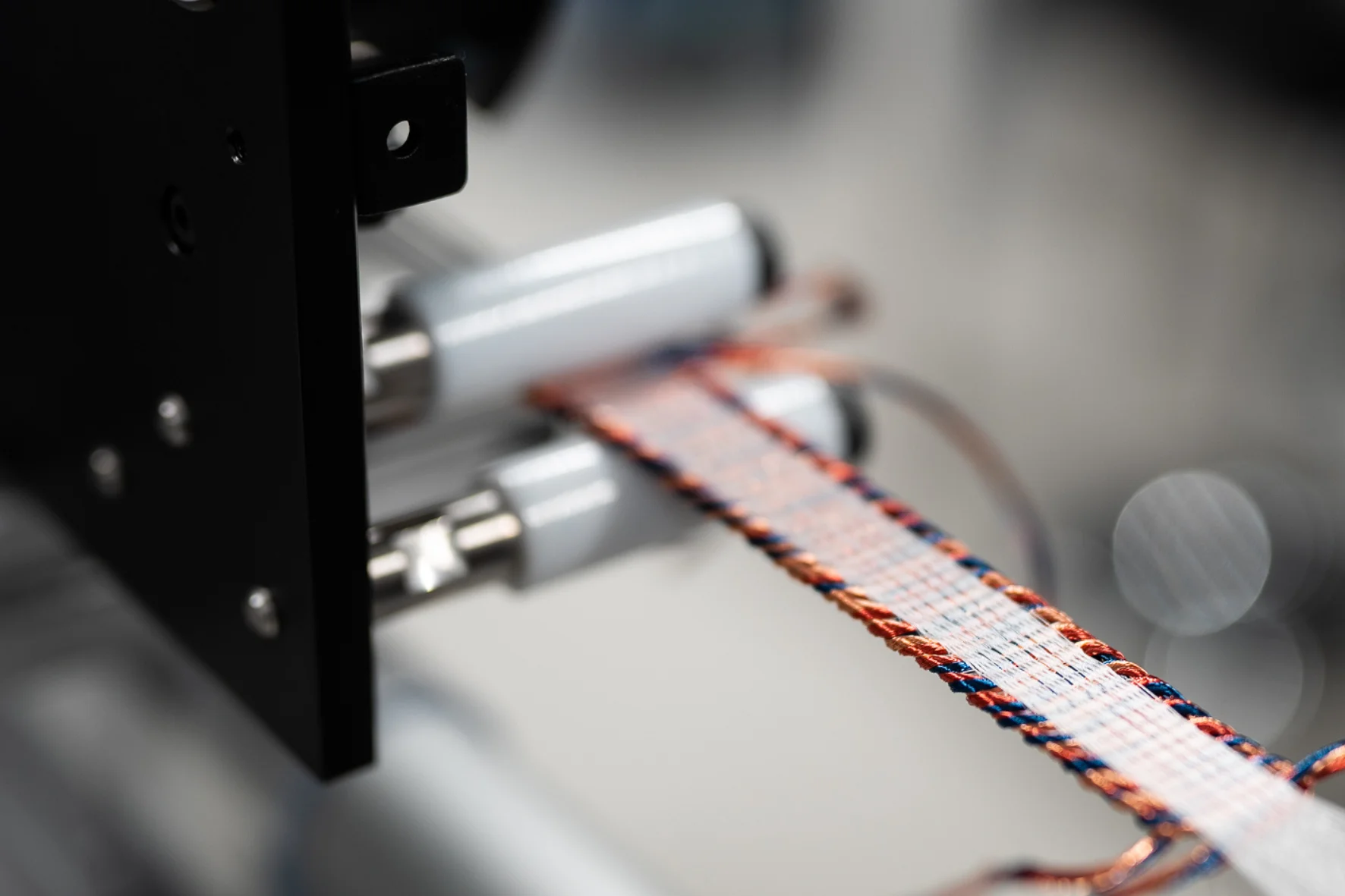 A close-up photograph of Alva Industries' manufacturing machinery, showing a flat, ribbon-like structure of woven copper and fiber strands being processed through precision rollers.