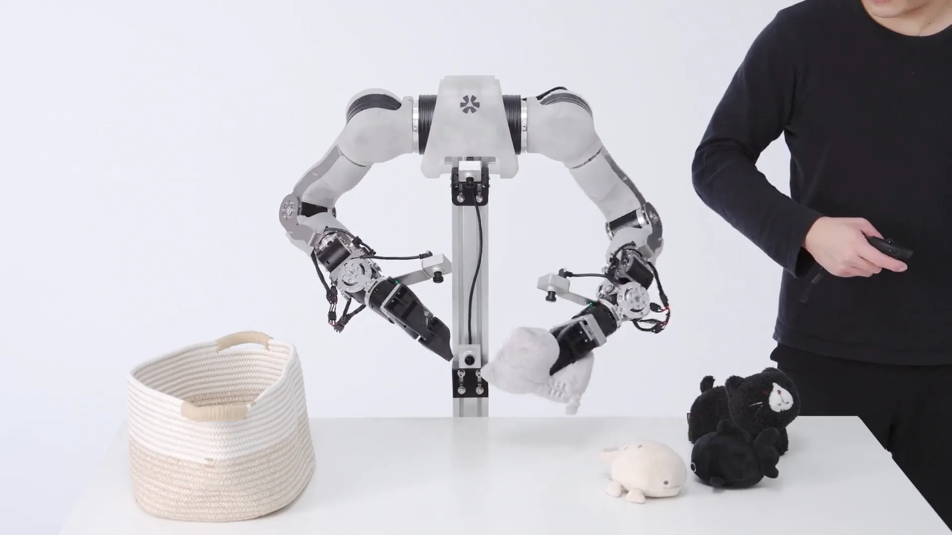 Anvil Robotics' OpenARM, an open-source bimanual manipulator, featuring two grey and black robotic arms mounted to a central vertical post. One arm is grasping a white plush toy above a white table, while a human operator to the right uses a handheld controller for teleoperation.