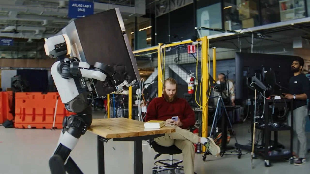 The all-electric Boston Dynamics Atlas humanoid robot balancing and carrying a black mini-fridge through a laboratory facility, walking past a seated engineer who is looking at a smartphone.