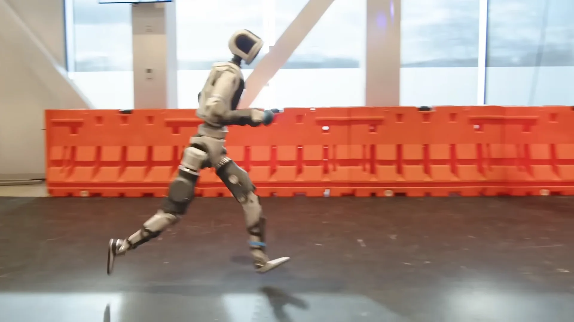 A side-profile view of the all-electric Boston Dynamics Atlas robot running in a laboratory environment. The robot is captured mid-stride with both feet clearly off the ground, demonstrating a dynamic flight phase, with orange safety barriers visible in the background.