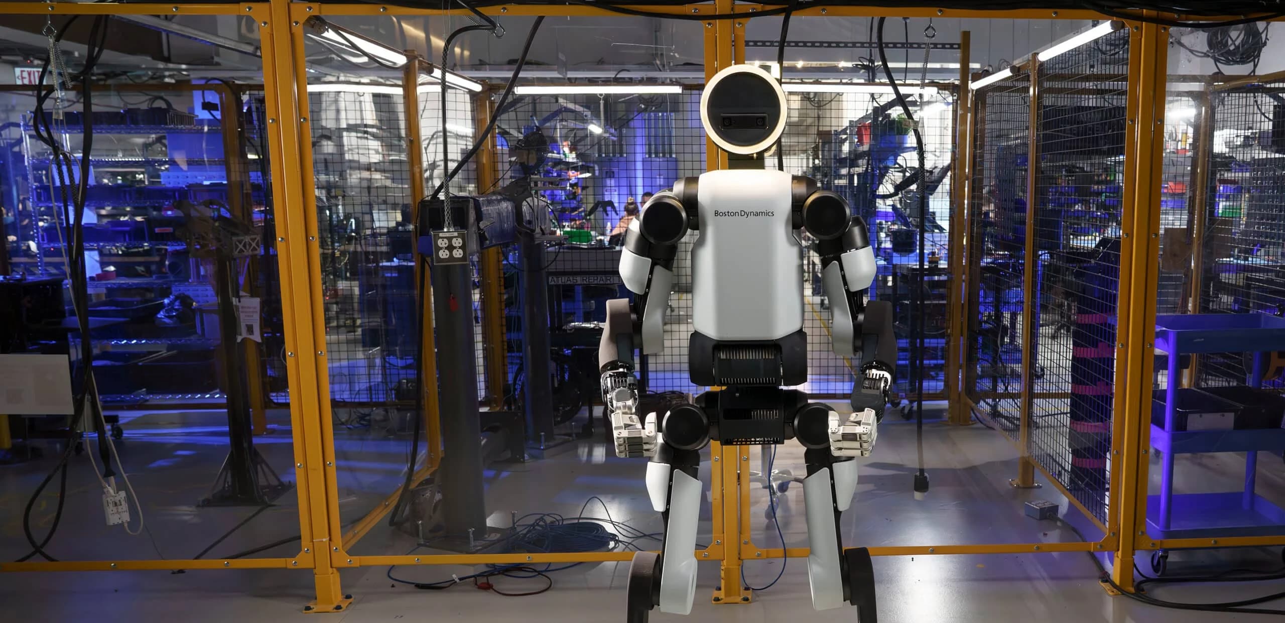 A full-length shot of the all-electric Boston Dynamics Atlas humanoid robot standing in a warehouse setting, centered behind yellow safety barriers. The robot's circular head is illuminated by a bright ring light.