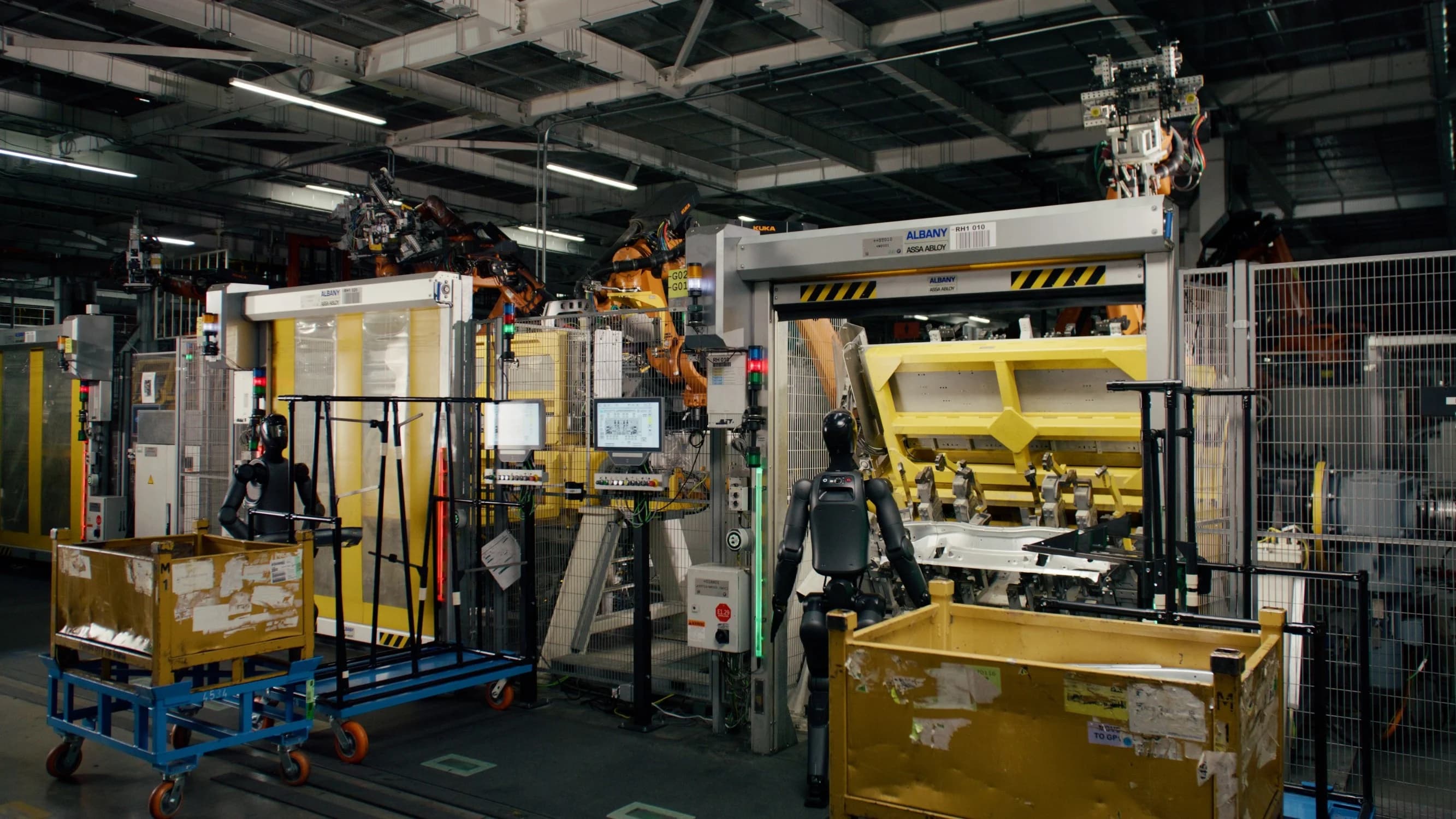 A wide industrial shot of two gray Figure 02 humanoid robots working in a large BMW body shop. The robots are surrounded by industrial safety fencing, yellow bins, and orange KUKA robotic arms.