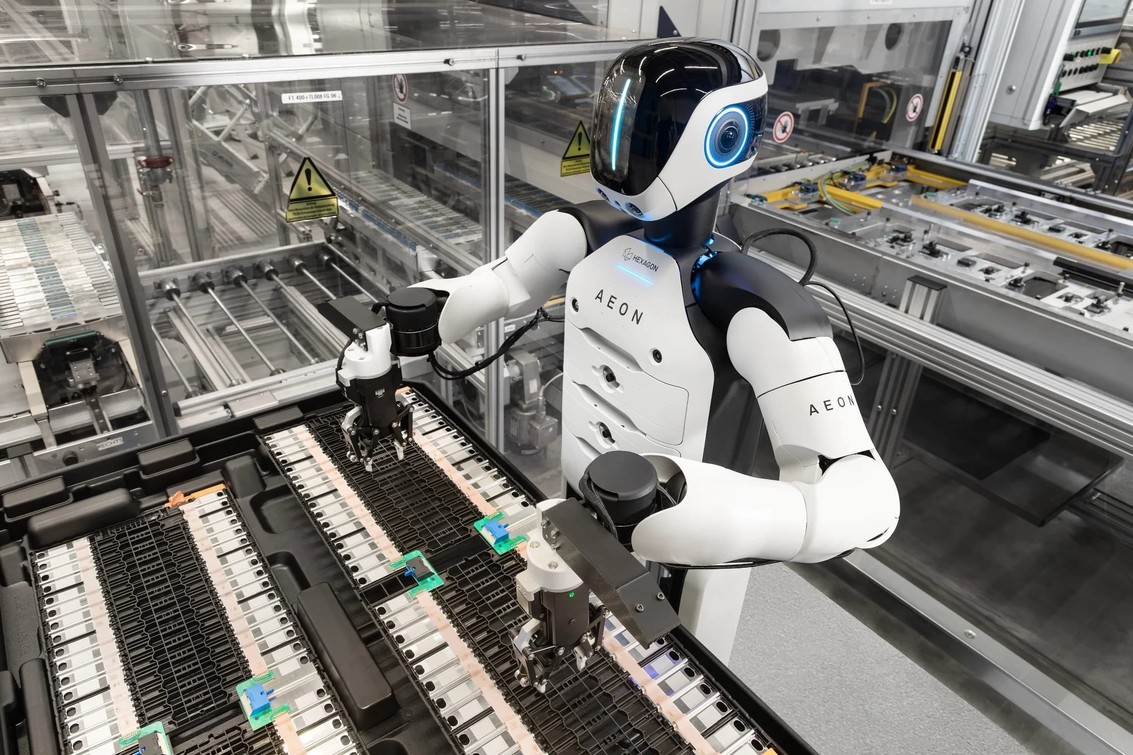 An angled shot of the AEON humanoid robot using high-precision robotic grippers to interact with rows of small electronic modules in a black tray. The robot's sleek white and black design is visible against a backdrop of industrial machinery.