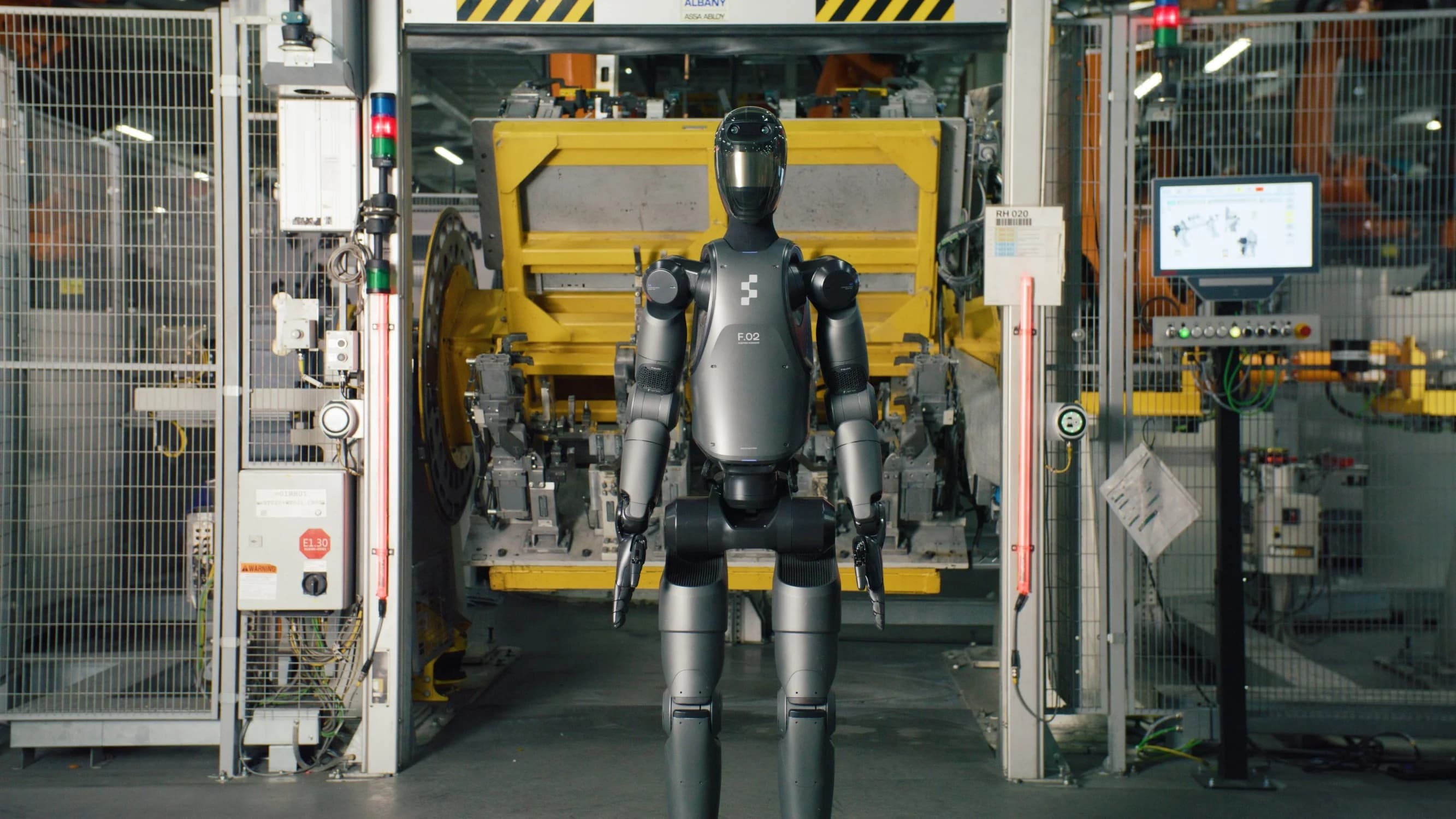 A front view of the gray Figure 02 humanoid robot standing in a BMW factory. The robot has 'F.02' printed on its chest and is positioned in front of a yellow industrial welding fixture.