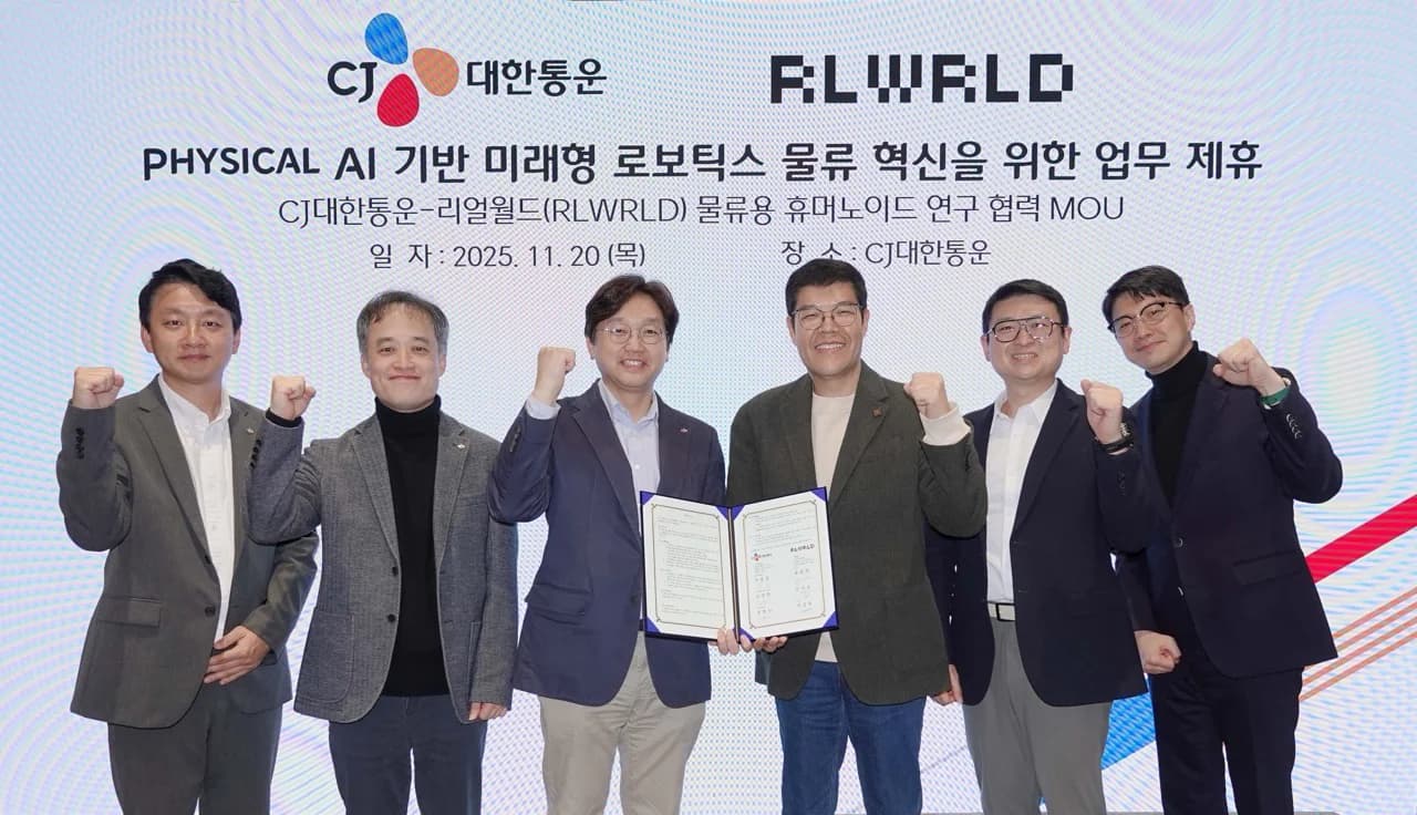 Six executives from CJ Logistics and RLWRLD pose for a group photo in front of a banner reading "Business Partnership for Future Robotics Logistics Innovation based on Physical AI." The two central figures hold a blue folder containing the signed agreement, while all six men raise their fists in a celebratory gesture.