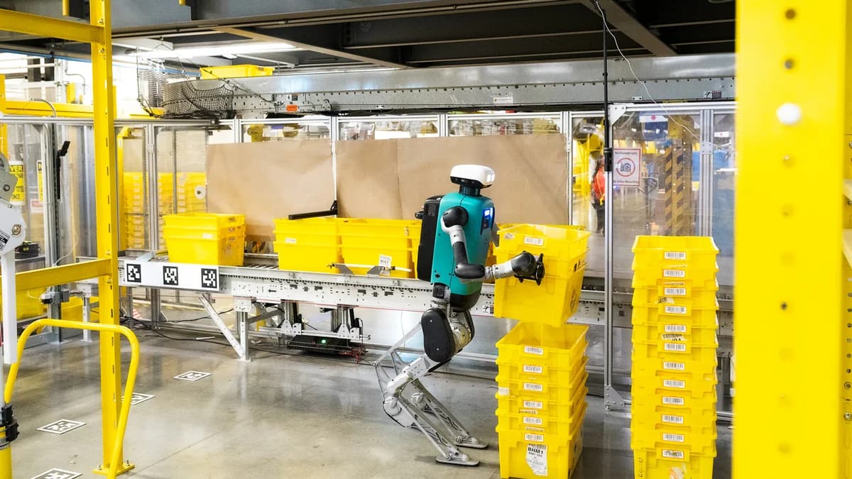 A full-body shot of Agility Robotics' Digit, a bipedal humanoid robot with a teal torso, lifting a yellow plastic tote in a warehouse setting. The robot stands on a concrete floor surrounded by yellow safety rails, stacks of bins, and conveyor belts marked with fiducial navigation tags.