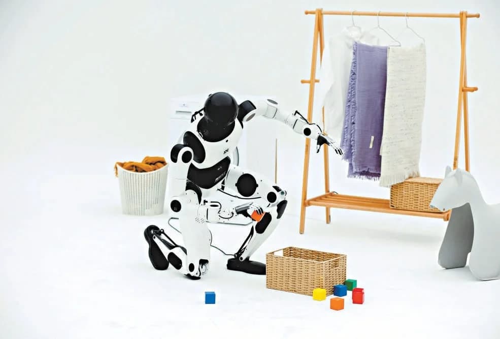 A black and white Embodied Tiangong 3.0 humanoid robot kneeling on a white surface to pick up colorful toy blocks. The robot is positioned in a household-like setting with a laundry basket, a wooden clothing rack with hanging fabrics, and a woven basket nearby.