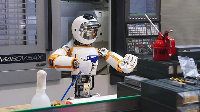 A white and orange humanoid robot with a digital visor displaying simple expressive eyes stands in an industrial machine shop. It is holding a blue compressed air nozzle with both hands, with a large CNC milling machine, a red oil can, and various workbench tools visible in the background.