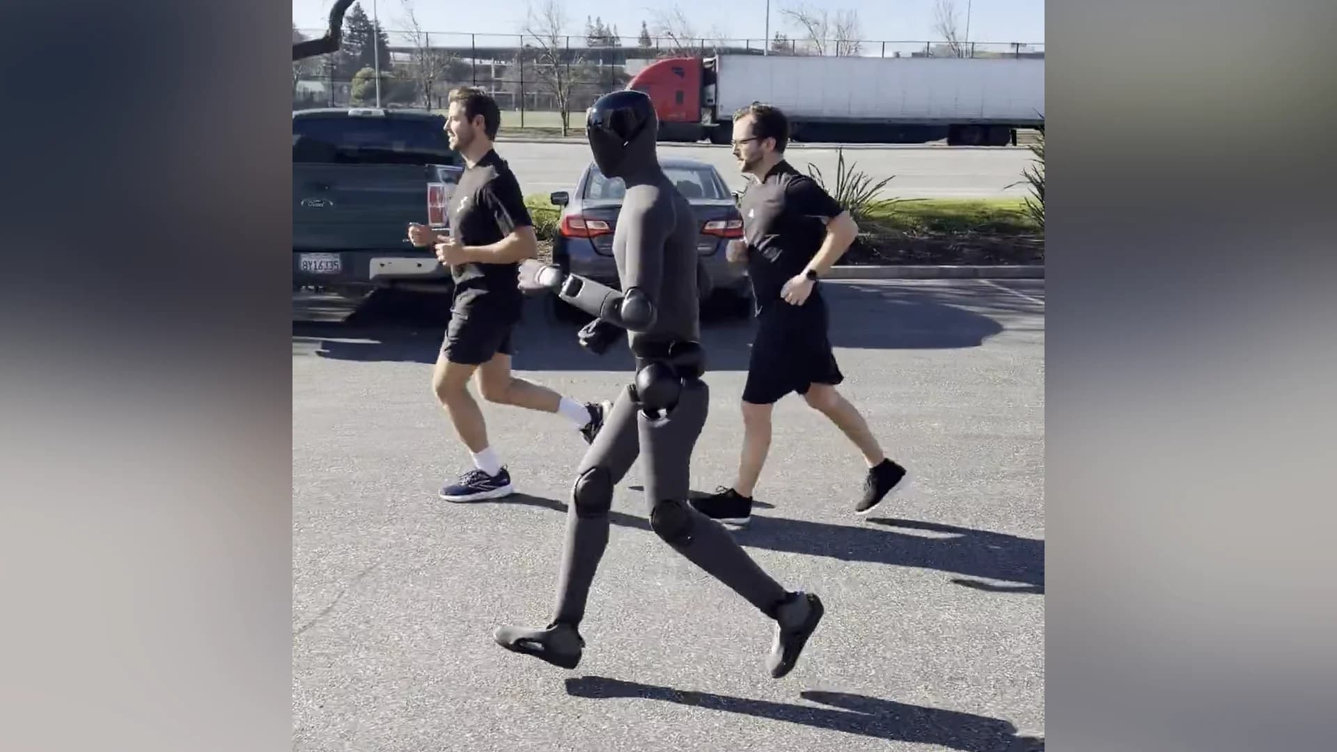 A dark gray Figure 03 humanoid robot jogging in a parking lot, flanked by two human runners.
