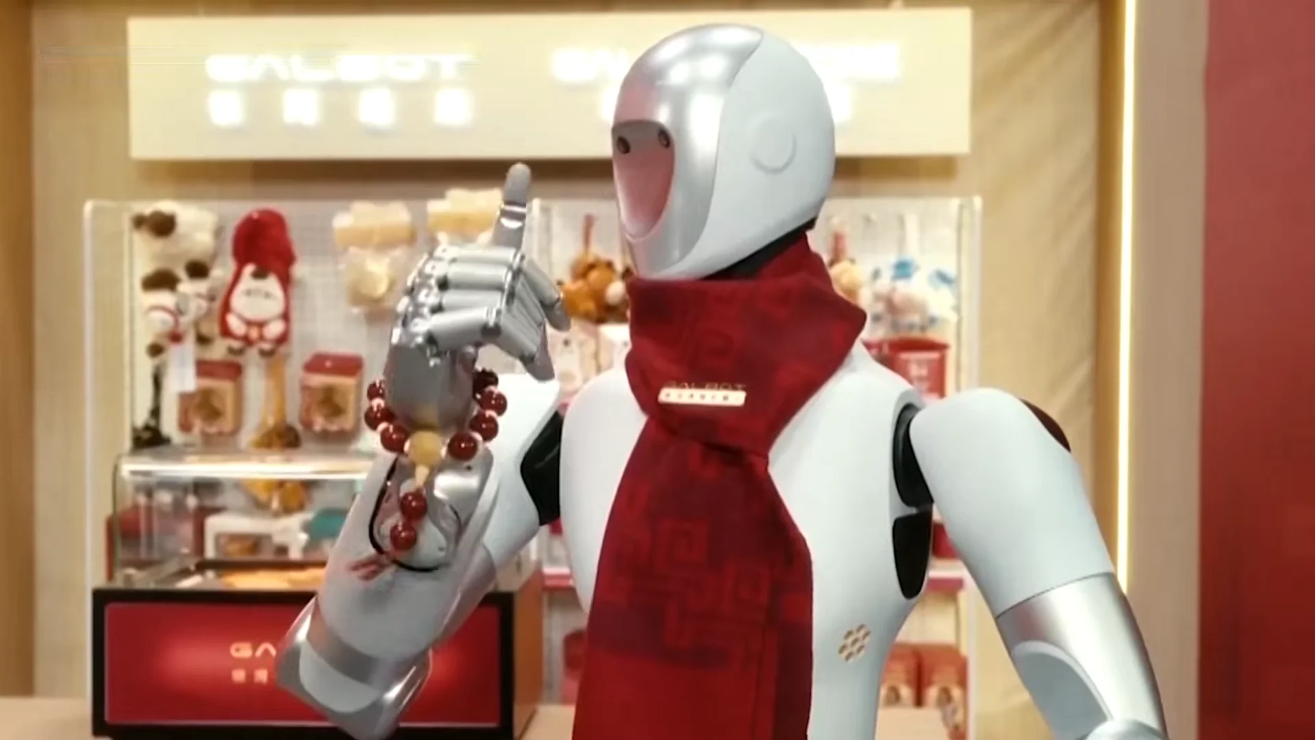 A silver and white Galbot humanoid robot wearing a red festive scarf and giving a thumbs-up gesture. It is holding a string of prayer beads in its right hand against a retail-like background.