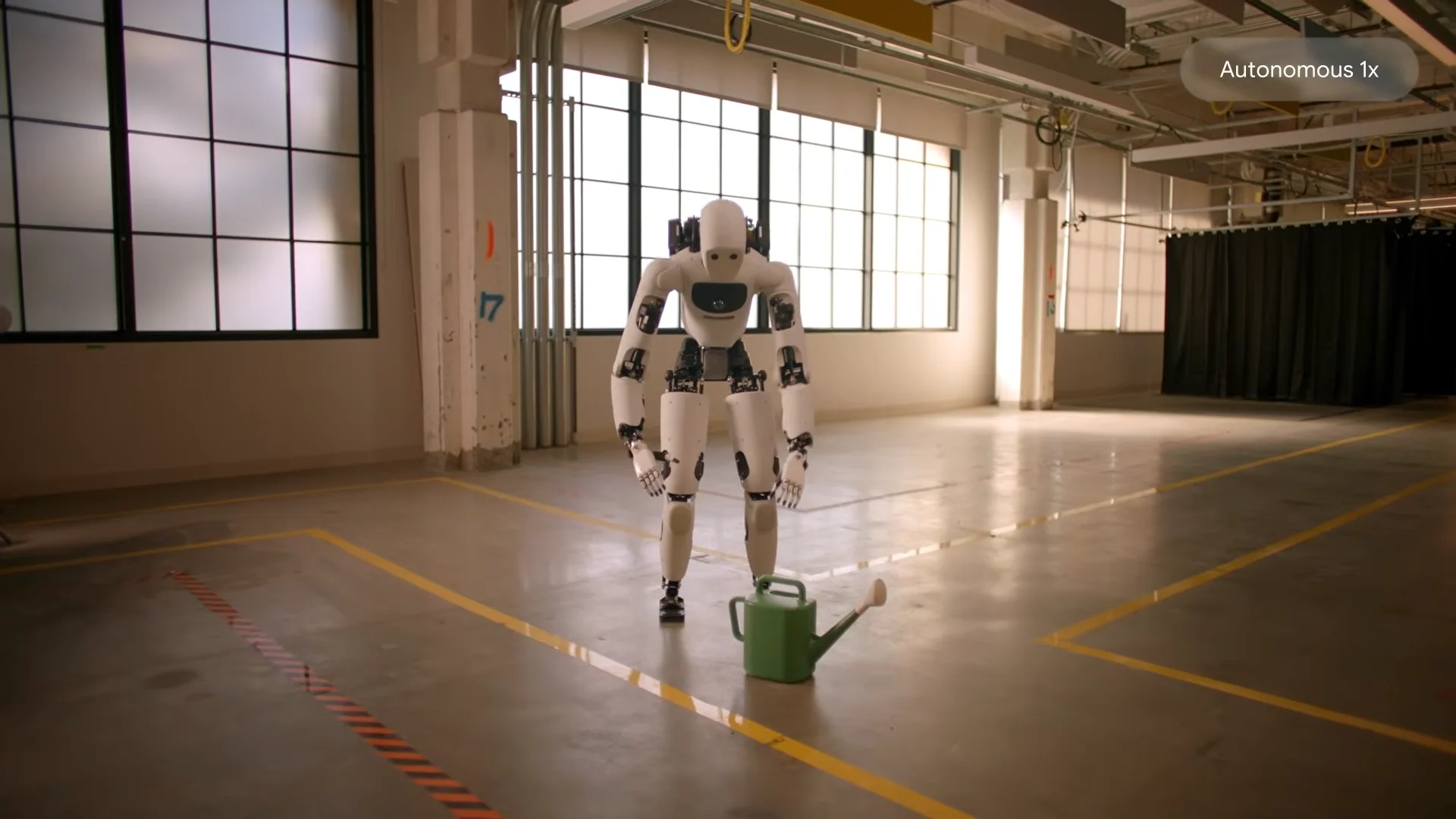 A white bipedal humanoid robot stands in a spacious industrial room, bending its knees and reaching down toward a green watering can on the floor. An overlay text reads 'Autonomous 1x'.