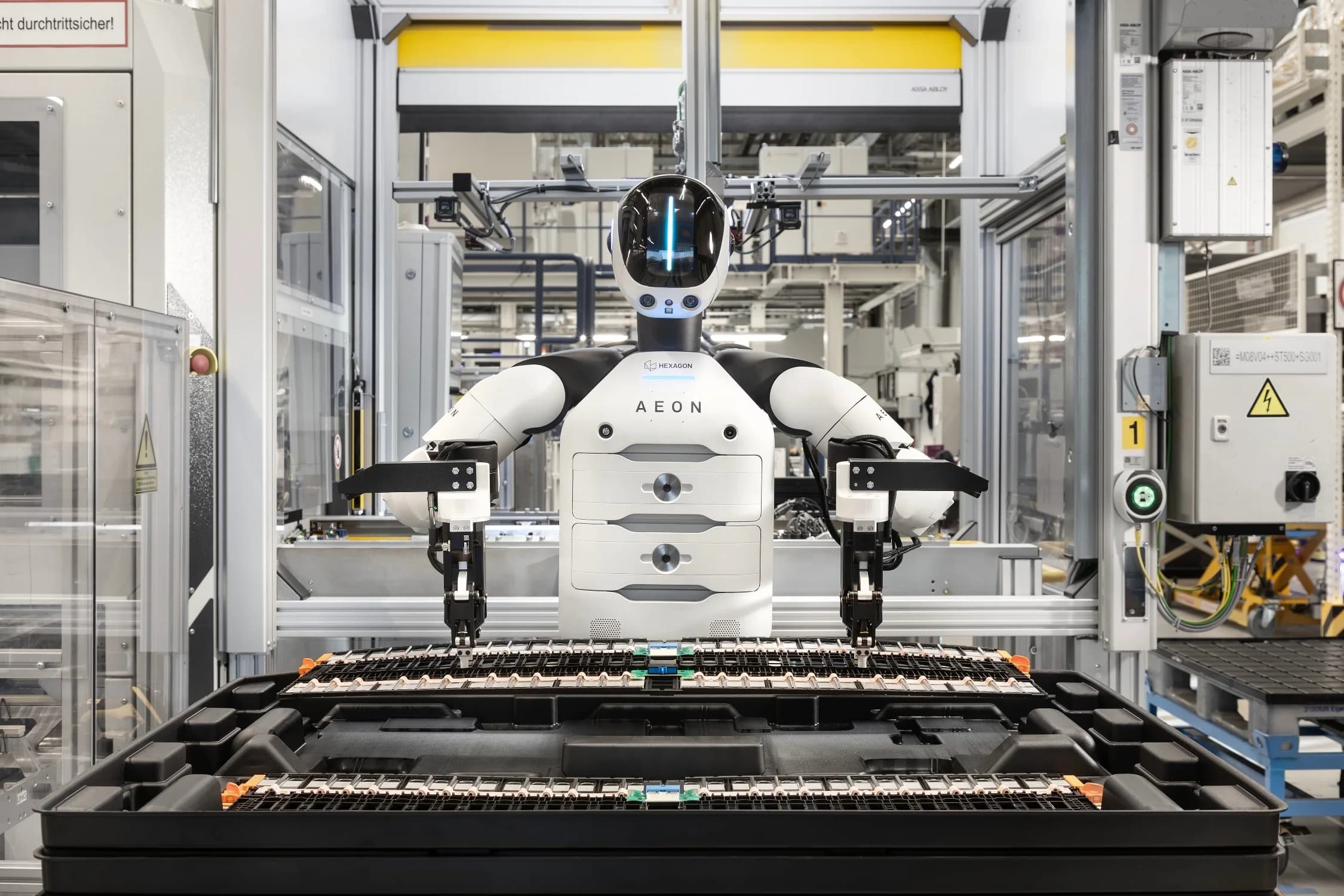 A front view of the white and black humanoid robot AEON, developed by Hexagon, standing behind a black tray filled with rows of small industrial components in a factory setting. The robot has two arms with specialized grippers positioned over the tray, and a sleek, black head with a glowing blue vertical light.