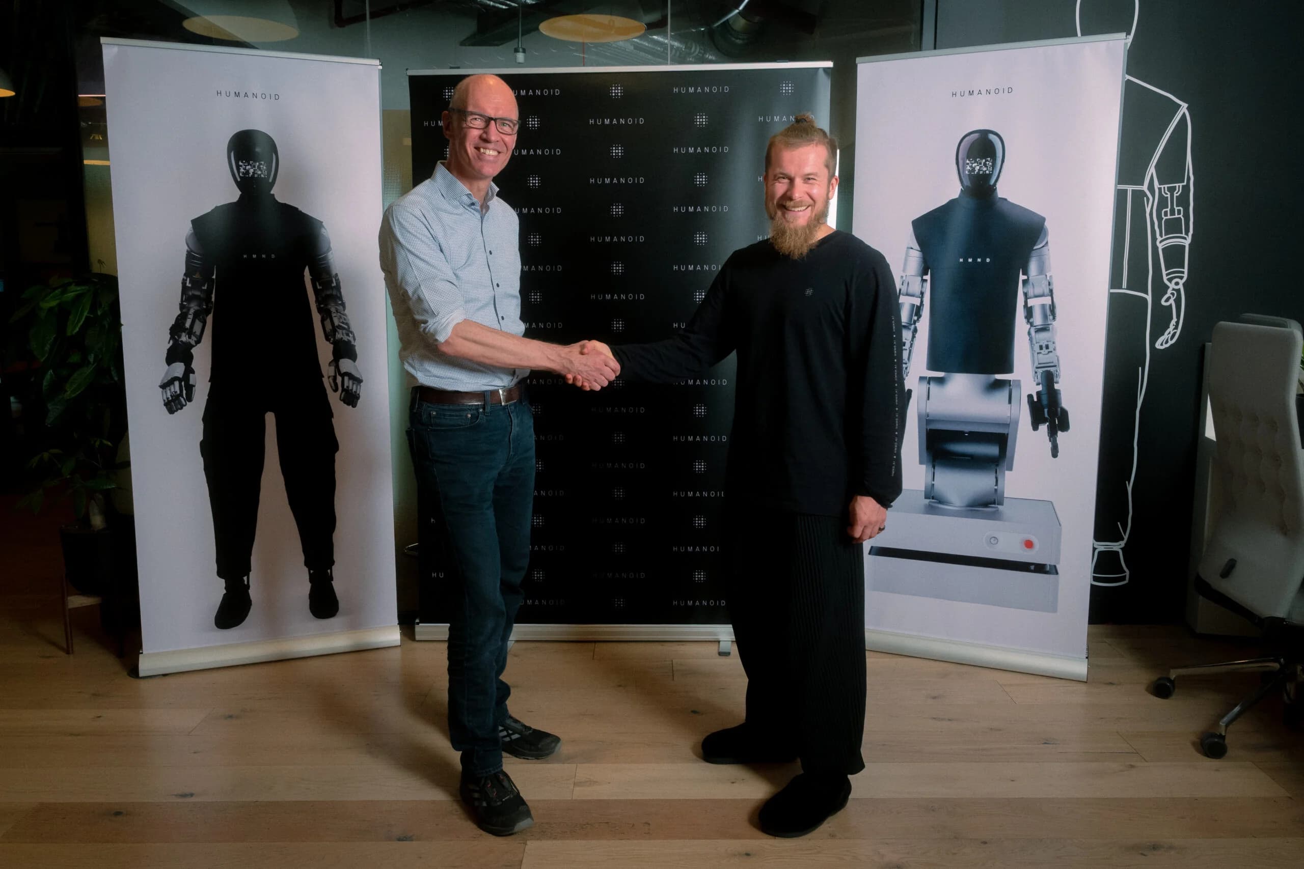 Two men stand smiling and shaking hands in an office space. The man on the left wears a light button-down shirt and jeans, while the man on the right wears an all-black outfit with a beard and hair tied back. Behind them are three tall banners: the center banner features the Humanoid logo repeated on a black background, while the flanking white banners display life-size images of the HMND 01 bipedal and wheeled robot variants.