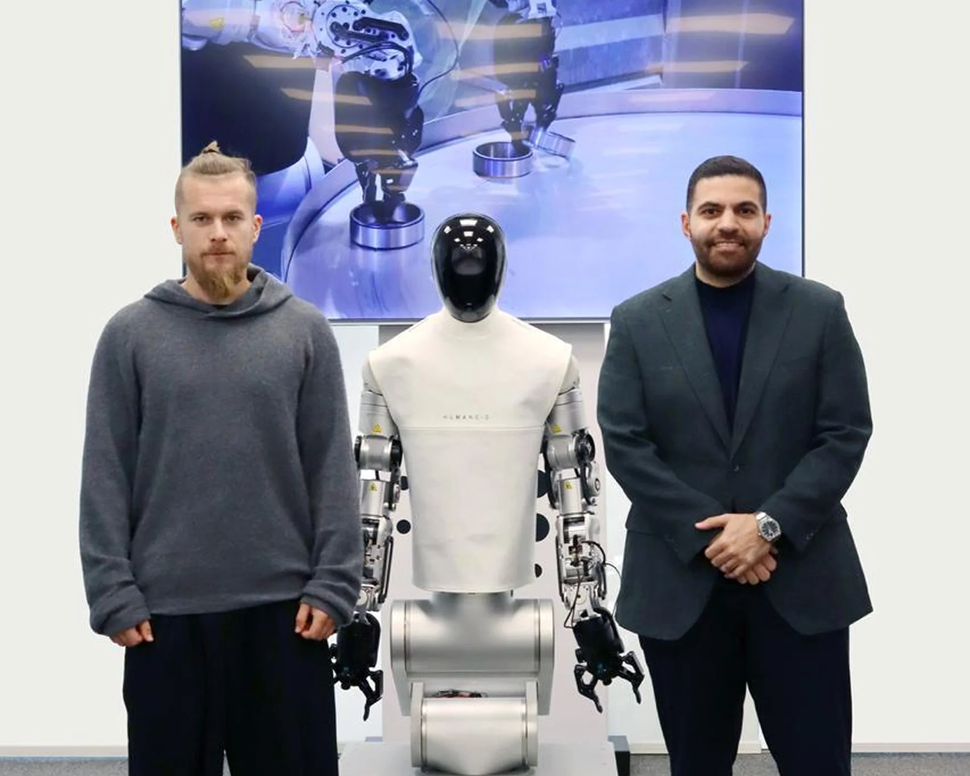 Artem Sokolov (left) and Dr. Elie Metri (right) stand on either side of the white and silver HMND-01 humanoid robot. A large screen in the background displays a close-up of the robot's hands performing a manipulation task.