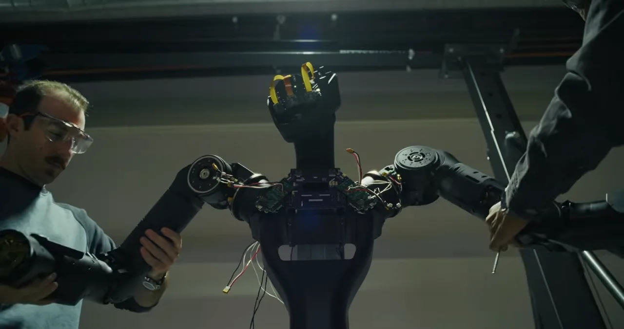 A low-angle shot showing the upper body assembly of a charcoal-black Foundation Phantom humanoid robot. Two technicians are visible, one on each side, manually attaching or adjusting the robot's arms. Internal electronics and wiring are exposed in the torso area.