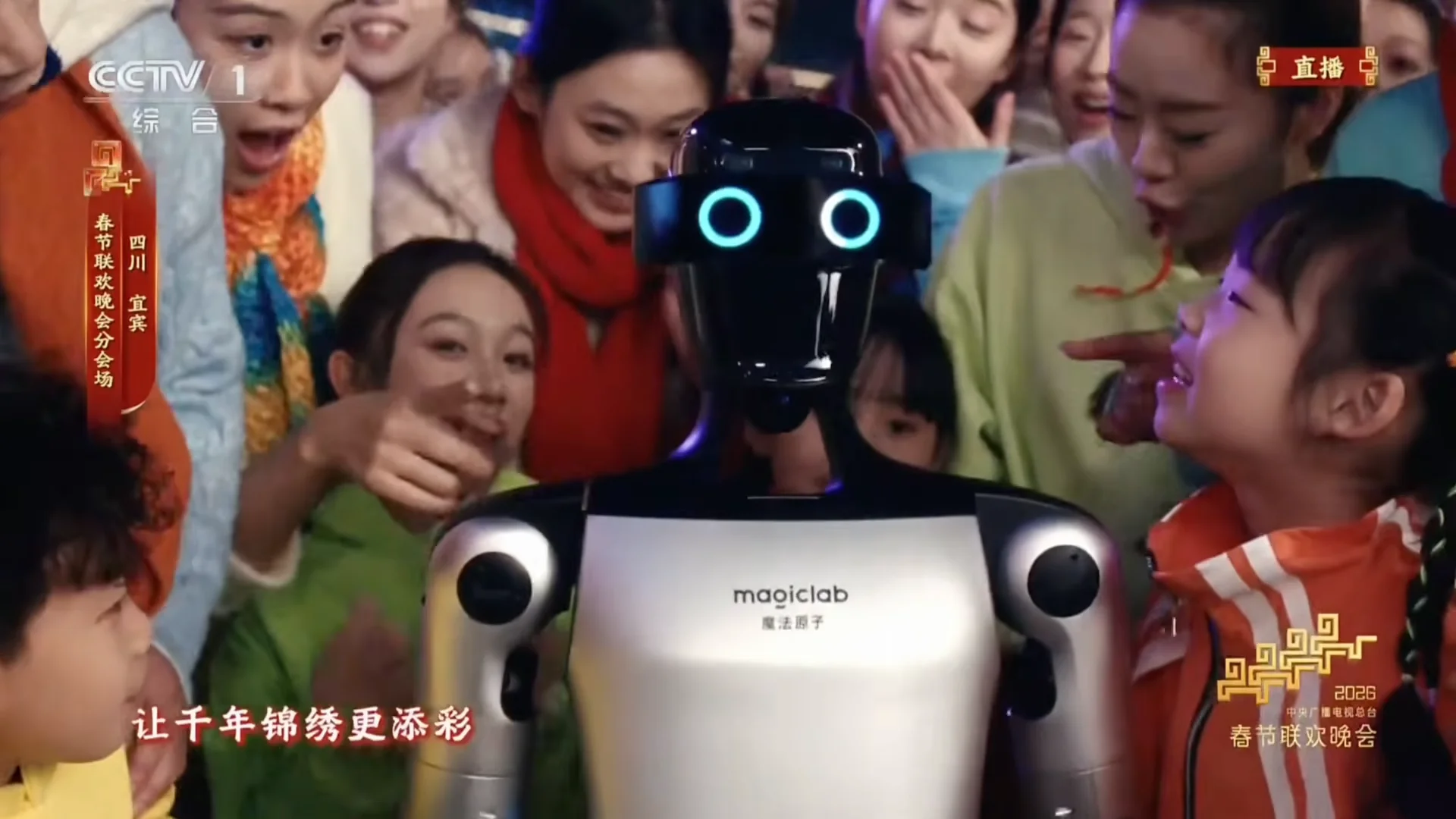 A close-up of a MagicLab humanoid robot with circular, glowing blue eyes and a silver-and-black torso. It is surrounded by a group of smiling children and adults. Logos for CCTV 1 and the 2026 Spring Festival Gala are visible in the corners.