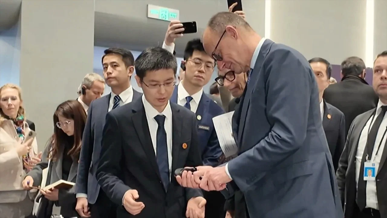 A close-up shot of Unitree founder Wang Xingxing holding a black mechanical robot component and explaining its function to German Chancellor Friedrich Merz. Merz is leaning in closely to examine the hardware while surrounded by members of a 30-person business delegation.