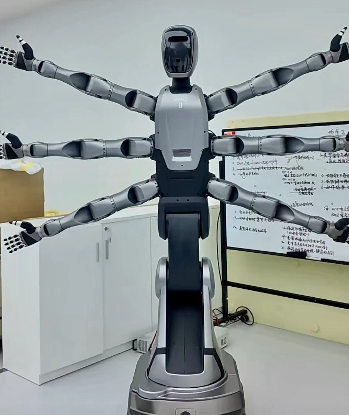 A studio shot of the Midea MIRO U robot standing in a laboratory. It has a silver humanoid head and torso, but features six bionic arms extending from its sides and a wheeled pedestal base instead of legs.
