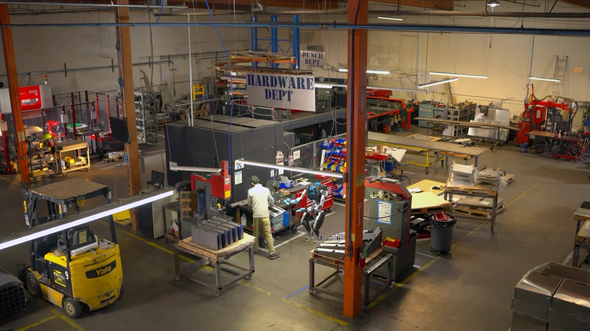 A high-angle view of an industrial workshop where a bipedal Noble Machines robot works side-by-side with a human employee in the hardware department.