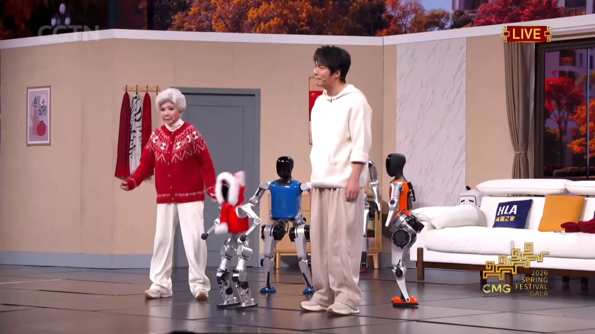 A scene from a comedy sketch during the 2026 Spring Festival Gala. On a stage designed to look like a living room, an older woman in a red patterned sweater and a younger man in a white hoodie stand together. A small silver humanoid robot wearing a red and white festive coat stands between them. In the background, two larger humanoid robots with blue and orange torsos are visible. The CGTN and 2026 Spring Festival Gala logos appear in the corners.