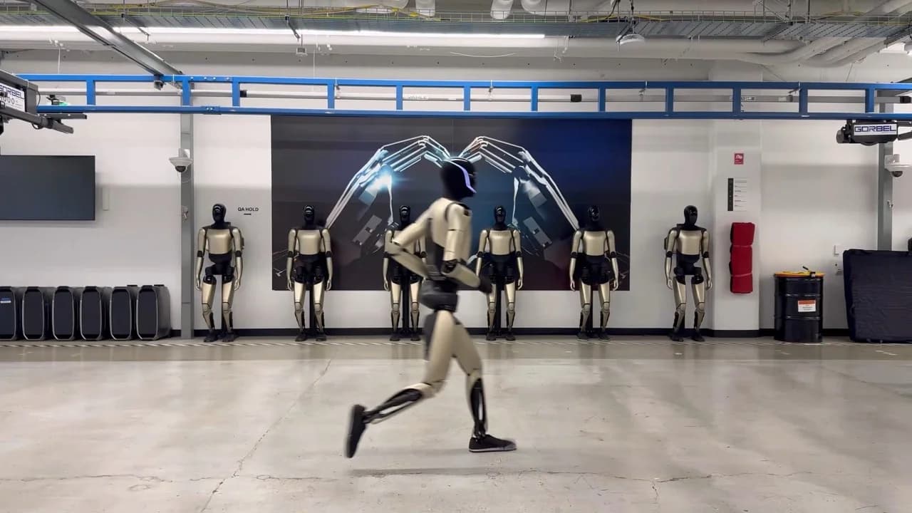 Side view of a Tesla Optimus humanoid robot sprinting across a concrete laboratory floor. The robot is captured in mid-stride with one leg lifted. In the background, a row of stationary Optimus units stands beneath a large wall graphic depicting two robotic hands forming a heart shape.