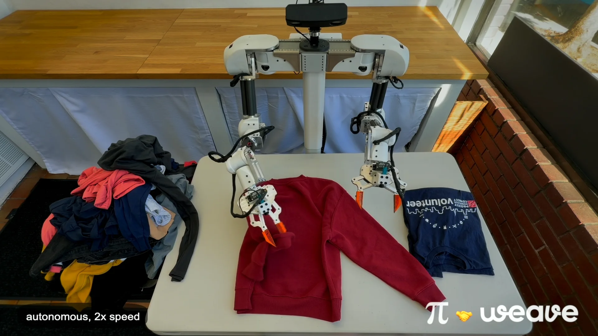 A high-angle view of the white Isaac 0 robot using its dual arms and three-finger grippers to fold a red sweatshirt on a white table. A pile of colorful laundry is visible to the left, while a neatly folded navy blue t-shirt sits to the right.