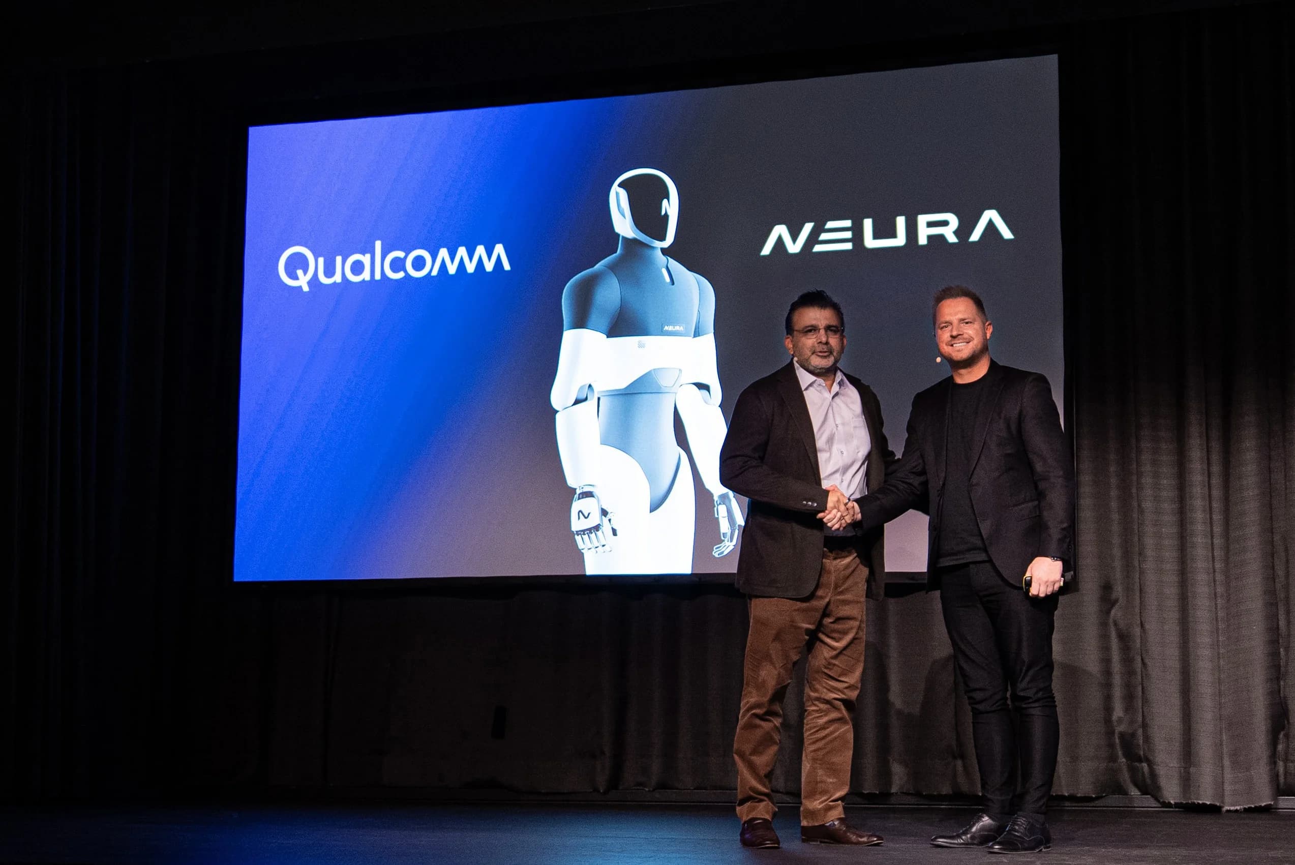 Nakul Duggal of Qualcomm and David Reger of NEURA Robotics shaking hands on a stage. Behind them, a large digital screen displays the Qualcomm and NEURA logos on either side of a rendering of a white-and-black humanoid robot.