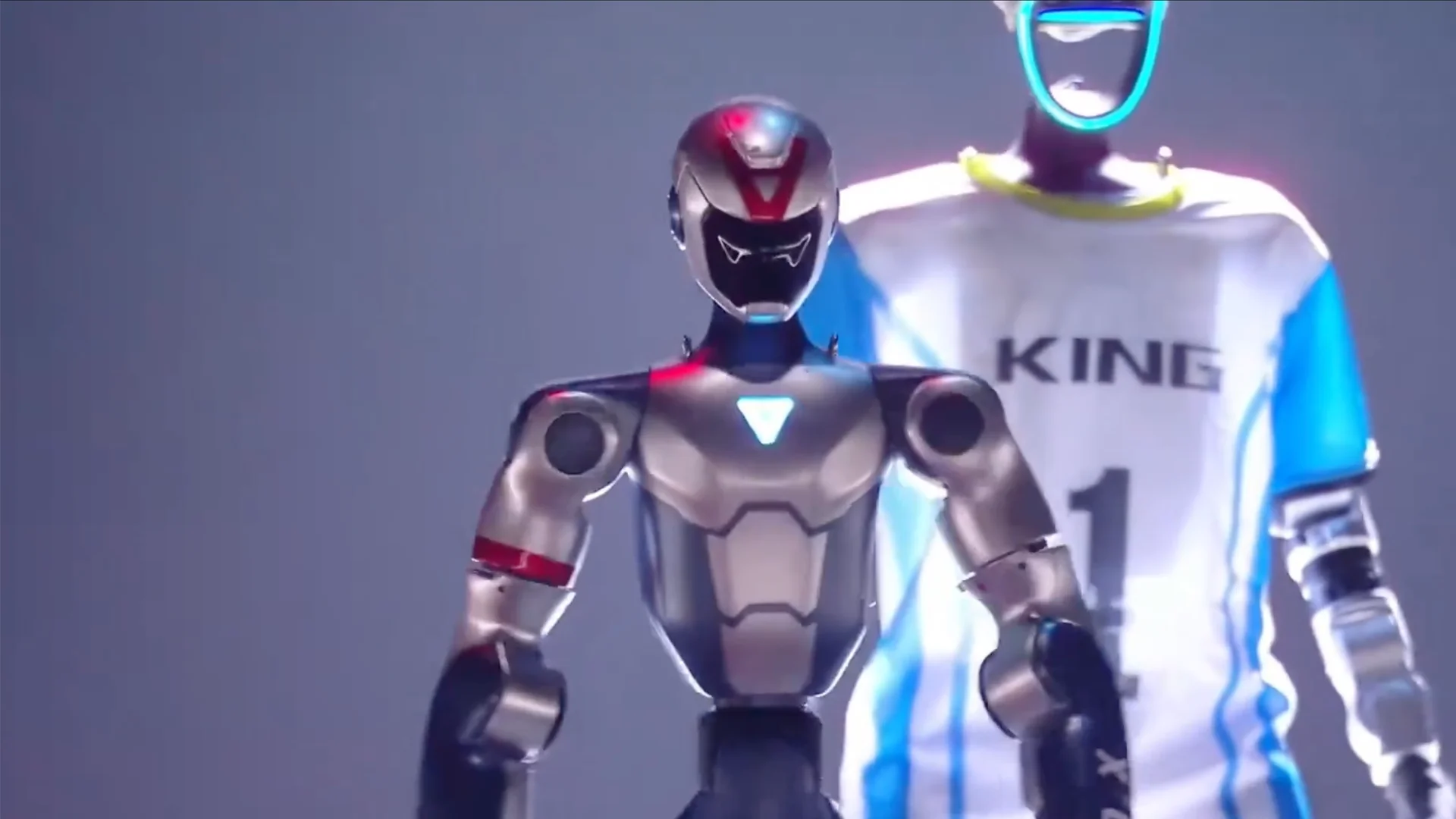 A close-up of a silver Unitree R1 humanoid robot standing in the foreground, with a taller Unitree G1 robot wearing a white and blue "KING" sports jersey standing behind it on a stage.
