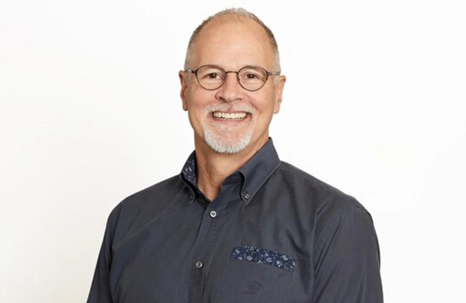 Headshot of Robert Playter smiling, with a grey goatee and glasses, wearing a dark blue button-down shirt with a patterned pocket square against a plain white background.