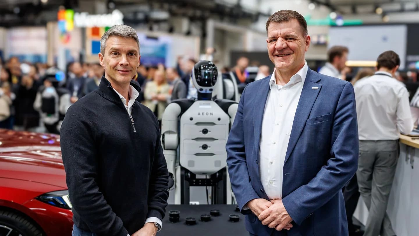 Arnaud Robert and Dr. Jochen Schröder stand on either side of the AEON humanoid robot at a crowded trade show. To the left, Robert wears a black quarter-zip sweater; to the right, Dr. Schröder wears a blue suit. The AEON robot between them has a white torso with black accents and a sleek black head. In the foreground, several small metallic robotic components are displayed on a black table.