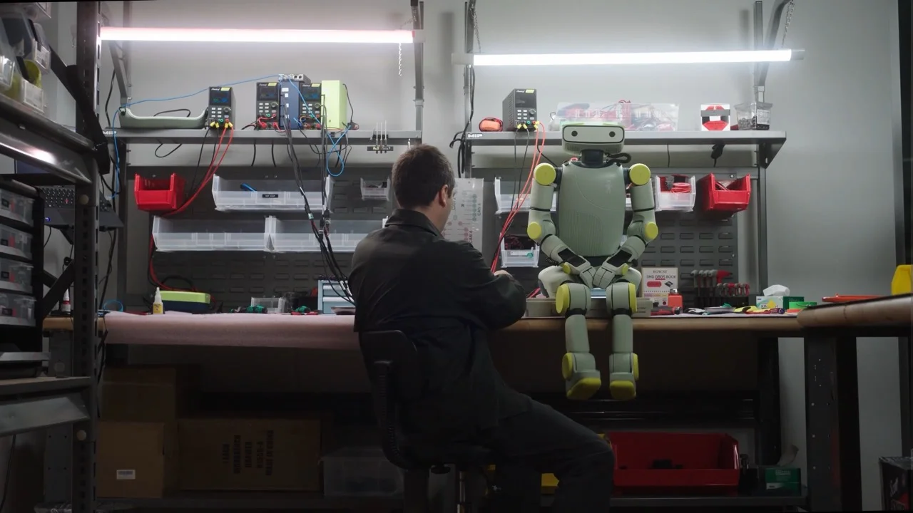A humanoid robot named Sprout, featuring a light green body and yellow joint accents, sits on the edge of a workbench. A person in a black jacket is seated in a chair, working on the robot. The background reveals a laboratory setting with power supplies, wires, and various tools on shelves.
