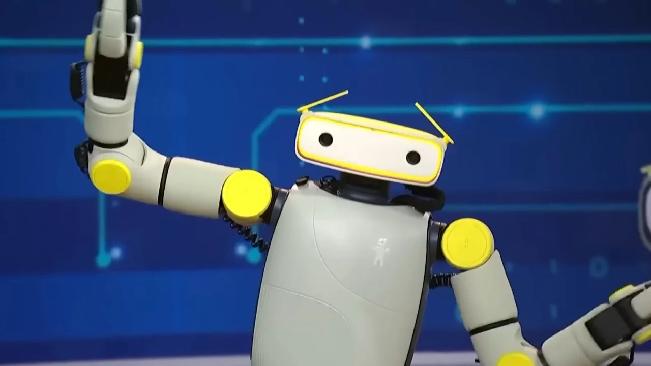 The Sprout humanoid robot celebrates in the NBC News studio with its arms raised. The robot features a light green torso, bright yellow shoulder and elbow joints, and a white rectangular head with two camera lenses and yellow, motorized eyebrows.