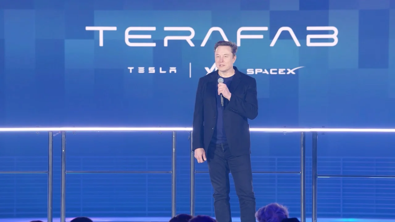 Elon Musk stands on a stage holding a microphone. Behind him, a large blue digital display features the word 'TERAFAB' in bold white text, with the logos for Tesla, xAI, and SpaceX listed beneath, separated by vertical lines.