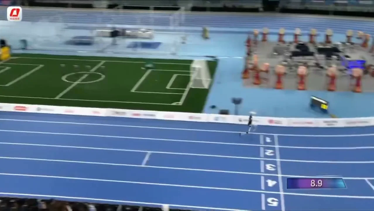 An elevated, wide view of a blue indoor running track where the Tiangong Ultra humanoid robot crosses the 100-meter finish line in lane one, with a broadcast on-screen timer displaying 8.9 seconds.