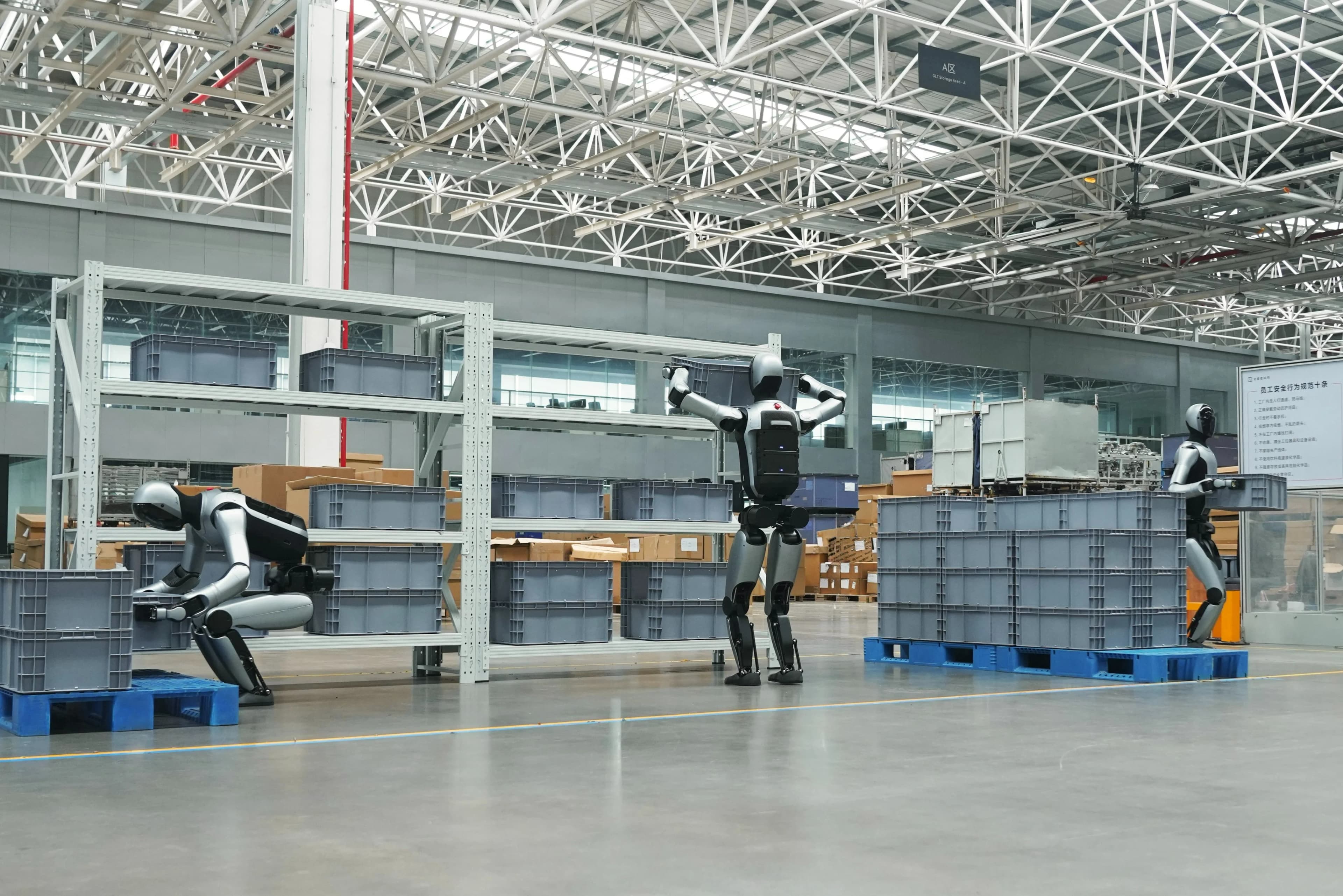 Three silver and black UBTECH Walker S2 humanoid robots working in a large industrial warehouse with high ceilings. On the left, one robot crouches to lift a gray crate from a pallet; in the center, another robot reaches up to place a crate on a high shelf; on the right, a third robot carries a crate across the floor.