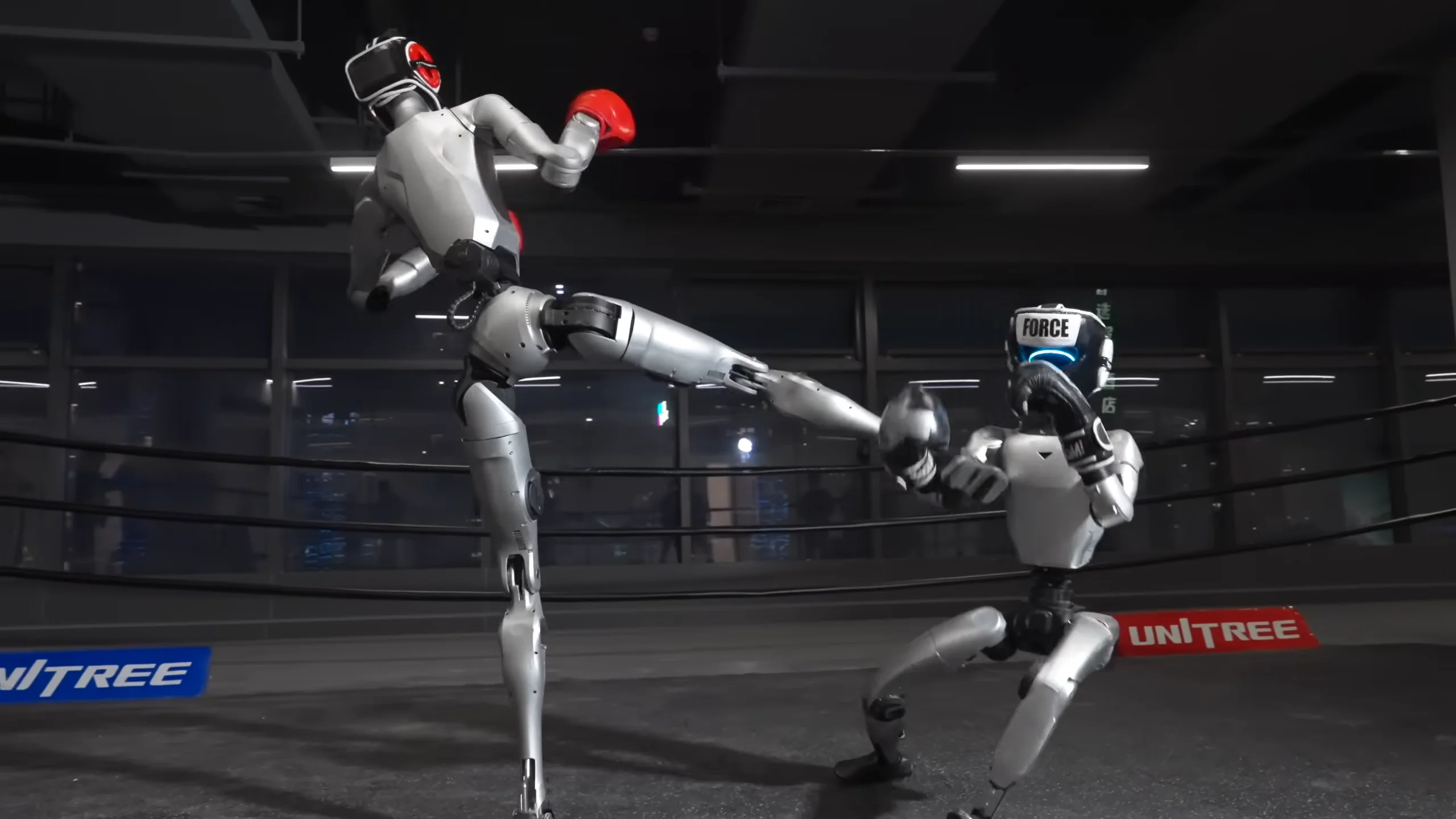 A screenshot from a Unitree promotional video showing the tall, silver H2 humanoid robot executing a high kick against the smaller G1 robot, which is crouching defensively in a boxing ring.