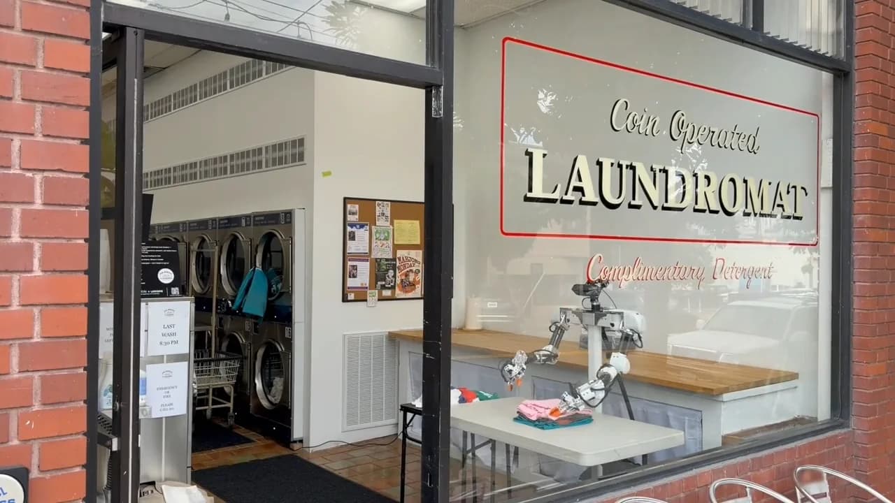 An Isaac robot prototype visible through a laundromat window, positioned at a folding table with stacks of laundry.