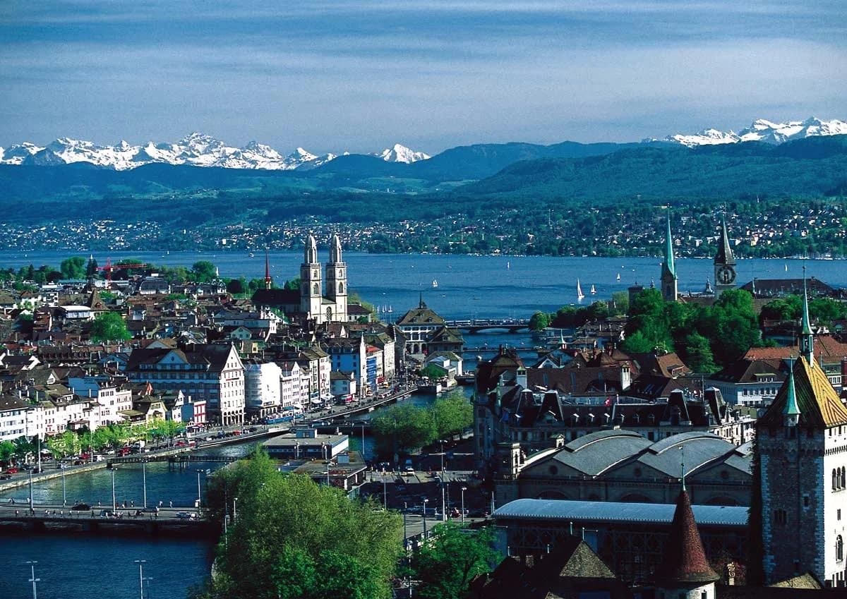 A scenic panoramic view of Zurich, Switzerland, featuring the Limmat River flowing through the historic city center, with the iconic Grossmünster twin towers and Fraumünster church visible. In the background, the snow-capped Swiss Alps rise sharply against a clear blue sky.